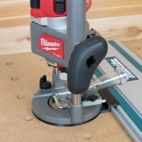 Milwaukee M18 2-1/4HP router mounted on gray plastic adapter base, positioned against Makita guide rail on wooden workbench