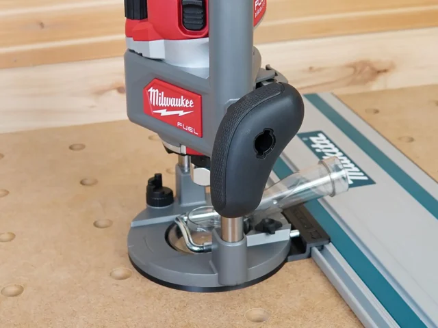 Milwaukee M18 2-1/4HP router mounted on gray plastic adapter base, positioned against Makita guide rail on wooden workbench
