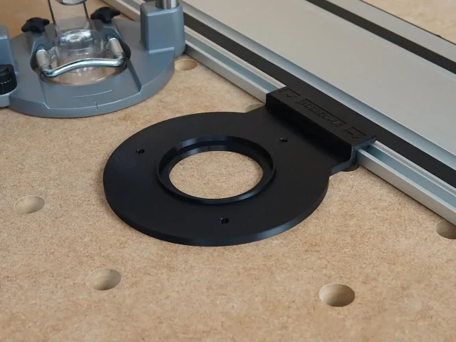 Black circular router adapter with central bore and mounting holes, positioned on pegboard next to Festool track rail