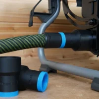 Black plastic hose adapter with blue band attached to green braided Festool hose, with a detached adapter shown on wood surface next to the tool