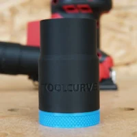Black ToolCurve hose adapter with blue textured grip ring, shown alone on wood surface with Milwaukee sander blurred in background