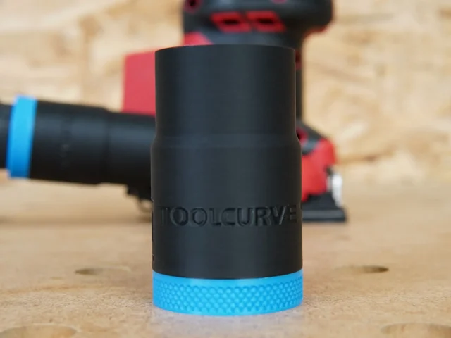 Black ToolCurve hose adapter with blue textured grip ring, shown alone on wood surface with Milwaukee sander blurred in background