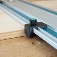 Black plastic drop stop mounted on Makita track saw rail with turquoise knurled knob, positioned at workpiece edge on cork base