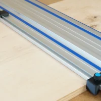Two black drop stops with blue knobs mounted on opposite ends of a Kreg track saw guide rail, positioned at the rail edges on a wooden workpiece