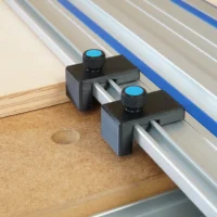 Two black drop stops with blue knurled knobs mounted in the slots of a Kreg ACS guide rail beside a plywood workpiece