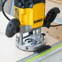 Black guide rail adapter bolted to a DeWalt DWE625 plunge router base, riding a green-edged track on plywood