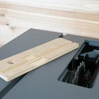 Light wood MDF zero clearance insert placed beside an open SawStop Compact table saw, showing the rectangular insert with a circular hole and slot detail