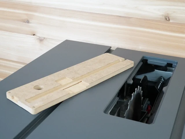 Light wood MDF zero clearance insert placed beside an open SawStop Compact table saw, showing the rectangular insert with a circular hole and slot detail