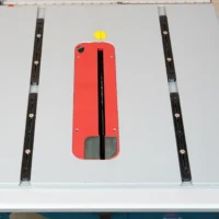 Red plastic miter bar with yellow knob and black slot, positioned centrally on a table saw surface with black adjustment bars on either side