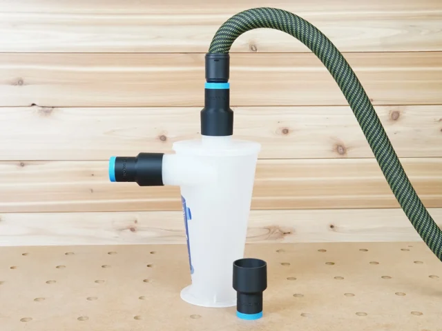 White plastic hose adapter with black threaded ports connected to green mesh hose and Oneida Dust Deputy, shown from front angle on wooden workbench