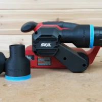 Black ToolCurve 2-1/2 inch hose adapter with blue trim, shown loose beside a SKIL belt sander with another adapter fitted to its dust port