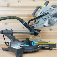 Black ToolCurve hose adapter with blue cuff joining a dust extractor hose to a Ryobi 10-inch sliding miter saw, spare adapter on the table