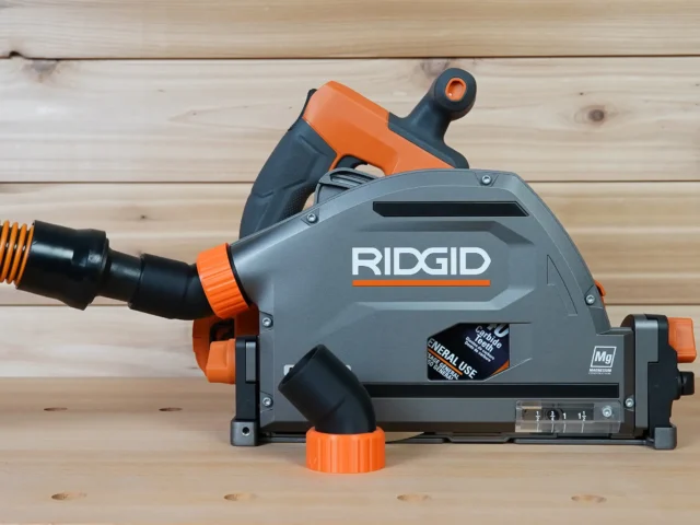 Black and orange no snag dust port adapter mounted on a Ridgid track saw, with orange-banded black vacuum hose attached to the upward-angled port