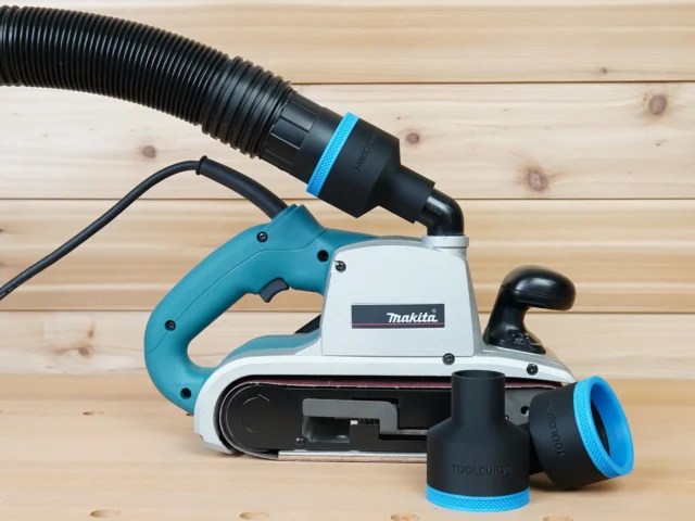 Black ToolCurve 2-1/2 inch hose adapter with blue ring joining a vacuum hose to a Makita 9403 belt sander, spare adapters beside it