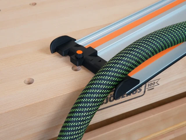 Black hose deflector mounted on a Wen track saw rail, with an orange quick-release clamping knob and green braided hose routed through the deflector