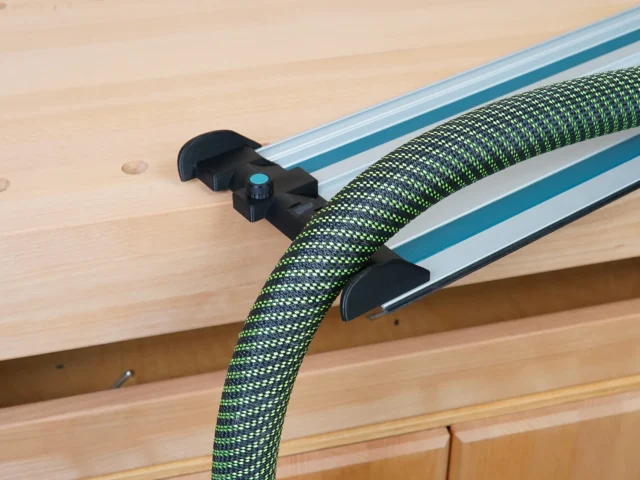 Black hose deflector mounted on Makita track saw guide rails, guiding green and black braided hose away from the cutting path