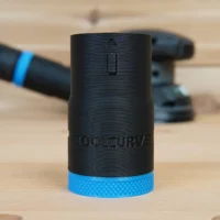 Black ToolCurve hose adapter with blue band, shown standalone on wood with a Flex sander and hose in the background