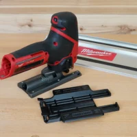 Milwaukee M12 Fuel jig saw with red and black adapter mounted on base, shown disassembled with track rail connector and replacement base plate on wooden workbench