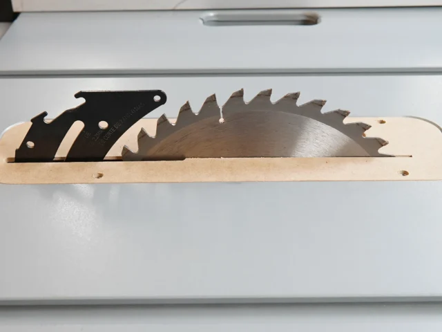 Wooden zero clearance insert with black metal blade holder and saw blade, displayed flat on white surface
