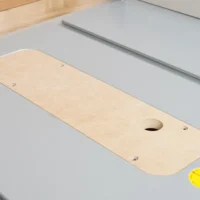 Natural wood MDF zero clearance insert with rounded ends and oval opening, mounted on gray table saw top with alignment holes visible