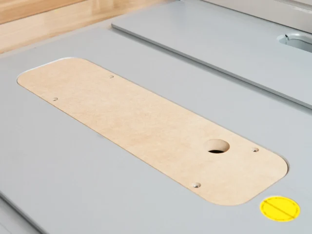 Natural wood MDF zero clearance insert with rounded ends and oval opening, mounted on gray table saw top with alignment holes visible