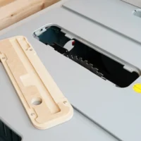 Light wood zero clearance insert for Bosch 4100 table saw, shown disassembled next to black plastic blade cover on gray saw surface