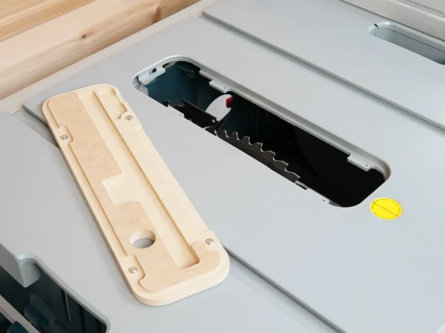 Light wood zero clearance insert for Bosch 4100 table saw, shown disassembled next to black plastic blade cover on gray saw surface