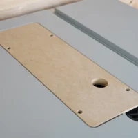 Tan MDF zero clearance insert with central blade slot and mounting holes, positioned on a Bosch table saw next to gray aluminum fence