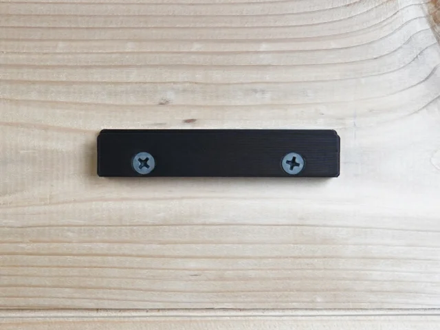 Black wall mount with two screw holes, designed to hold a track saw guide rail square vertically on a workbench or cabinet