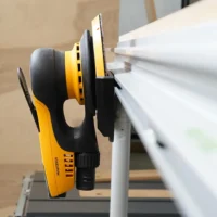 Yellow and black sander holder mounted to white MFT/3 table rail, cradling a random orbital sander with dust extraction hose