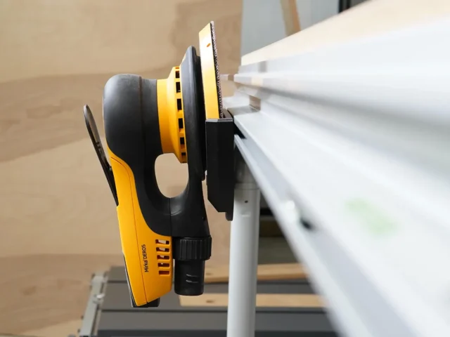 Yellow and black sander holder mounted to white MFT/3 table rail, cradling a random orbital sander with dust extraction hose