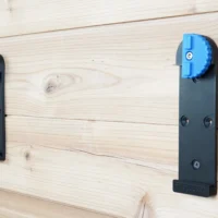 Two black wall mounts with blue locking clamps installed on light wooden boards, showing different sizes and mounting hole patterns