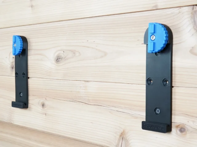 Two black wall mounts with blue locking clamps installed on light wooden boards, showing different sizes and mounting hole patterns