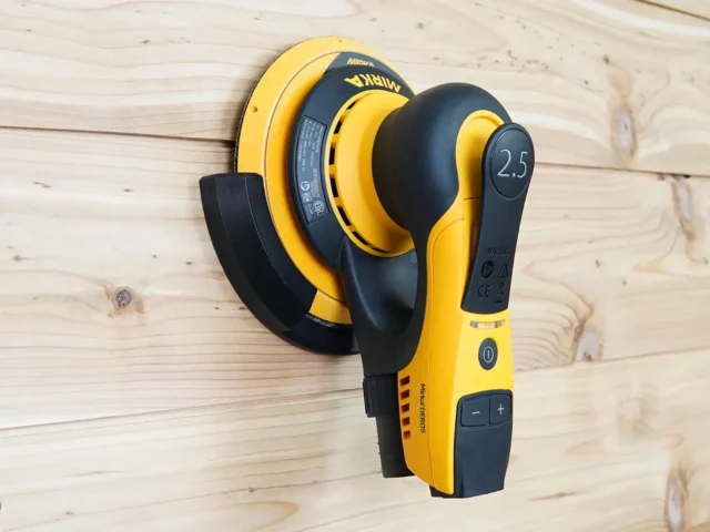 Yellow Mirka DEROS sander resting in a black wall mount holder screwed to a pine plank wall
