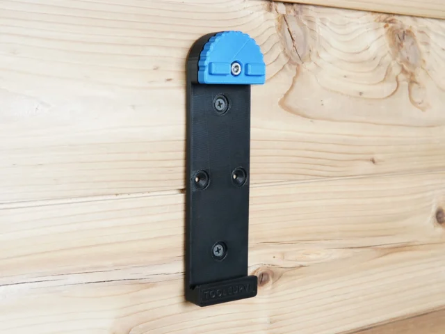 Black wall mount with blue top clamp attached to wooden wall, showing three mounting holes and ToolCurve branding