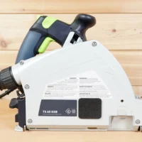 White dust cover with warning labels and Kick Back Stop logo installed on Festool TS 60 track saw, front view showing arbor opening coverage