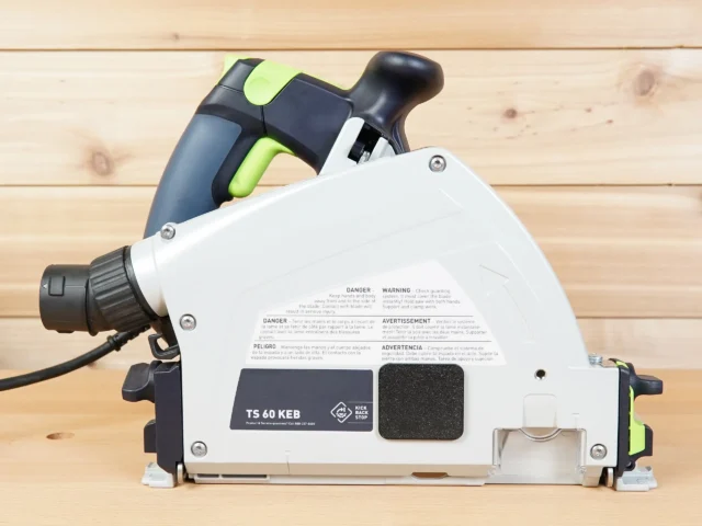 White dust cover with warning labels and Kick Back Stop logo installed on Festool TS 60 track saw, front view showing arbor opening coverage