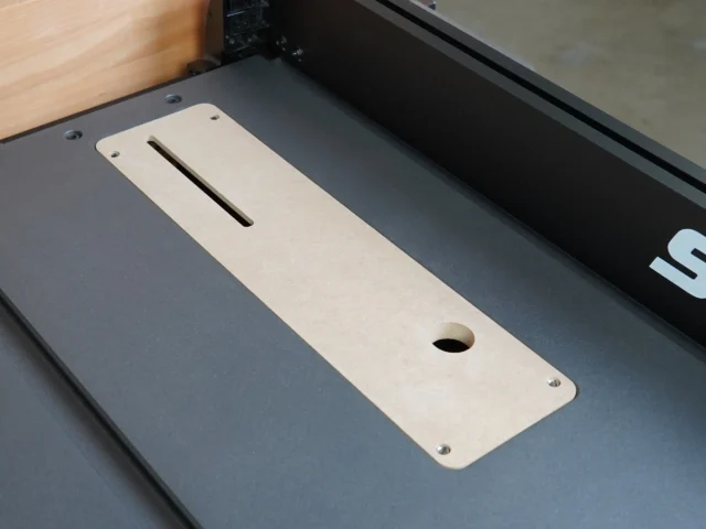 Light wood MDF zero clearance insert with centered blade slot and round hole, mounted on dark table saw bed with mounting screws visible