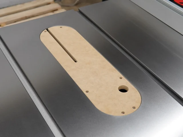 Light wood MDF zero clearance insert with rounded ends, center slot, and mounting holes, positioned on a gray table saw insert opening
