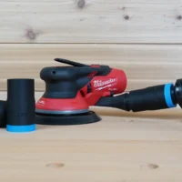 Black 1-1/4" hose adapter with blue trim linking an orange hose to a Milwaukee M12 sander, spare parts beside