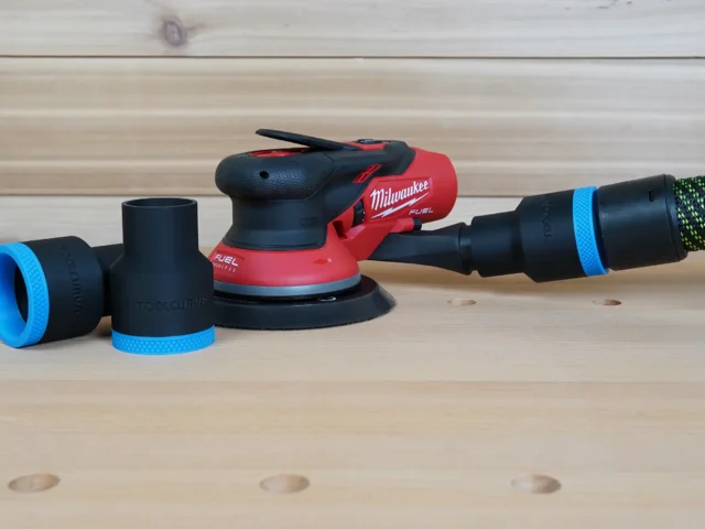 Black and blue 36mm hose adapter connecting a vacuum hose to a Milwaukee M12 Fuel sander, with two spare adapters beside it
