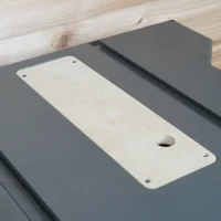 White MDF zero clearance insert with center blade slot and screw holes, positioned on a dark gray table saw top