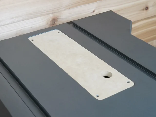 White MDF zero clearance insert with center blade slot and screw holes, positioned on a dark gray table saw top