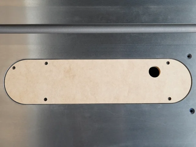 Light MDF zero clearance insert with rounded ends, four mounting holes, and one blade slot, positioned on a gray metal table saw surface
