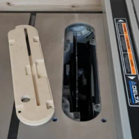 Cream-colored MDF zero clearance insert for Delta table saw alongside black factory insert, showing slots and mounting holes
