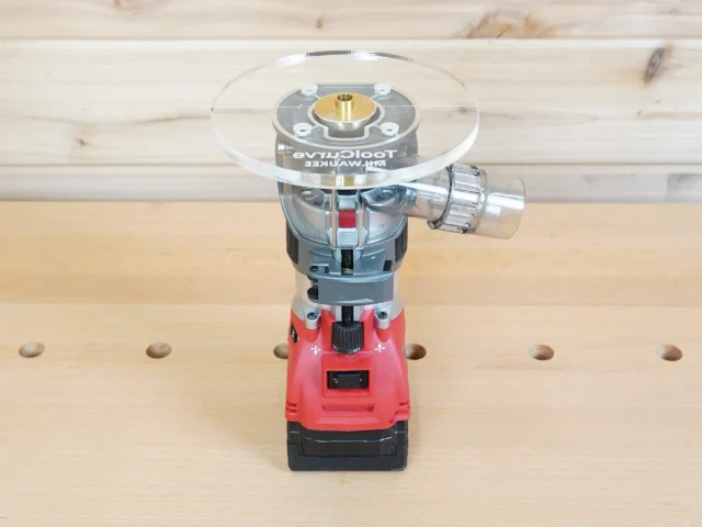 6-inch clear acrylic router base plate mounted on red Milwaukee trim router, shown from above on workbench with pegboard behind