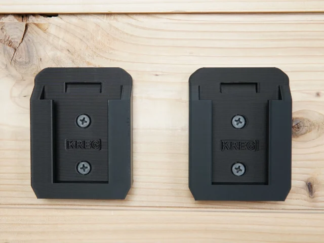 Two black plastic battery mounts for Kreg 18v batteries mounted on light wood, each with a central slot and two screw holes
