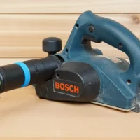ToolCurve hose adapter attached to a blue Bosch planer, connecting a black and blue Festool dust extractor hose to the tool's outlet port
