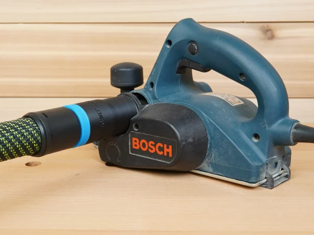 ToolCurve hose adapter attached to a blue Bosch planer, connecting a black and blue Festool dust extractor hose to the tool's outlet port