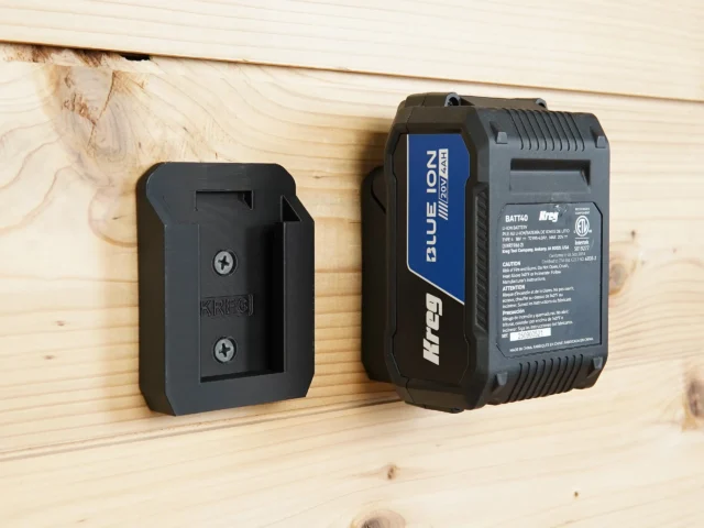 Black Kreg battery mount attached to wood wall next to a Kreg 18v Blue Lion battery, showing slot for battery to click into place